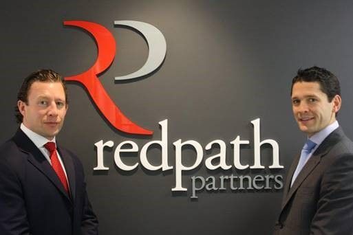 Two professional men in suits stand in front of a Redpath Partners logo on a grey office wall.