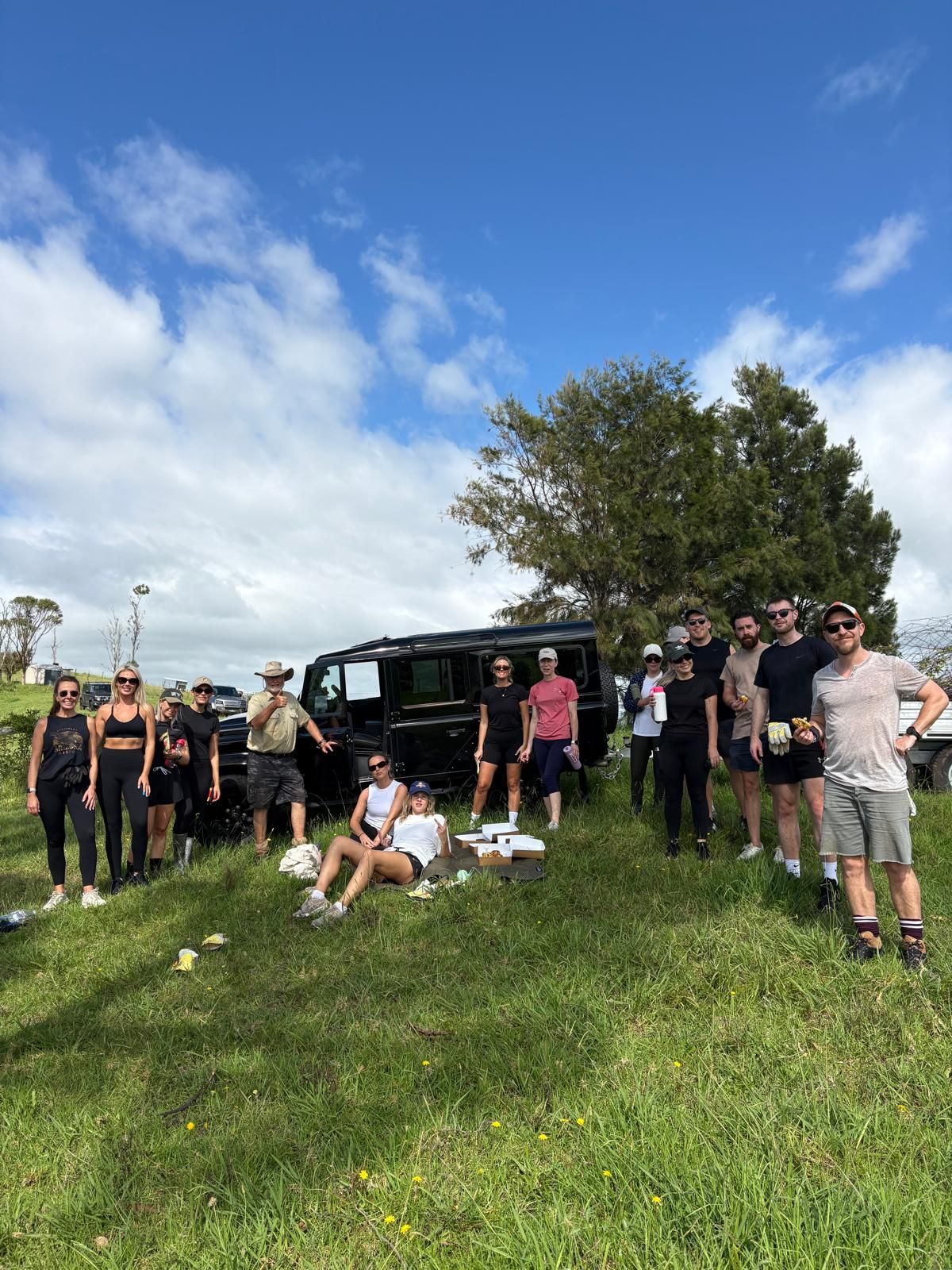 A group of people standing and sitting on grass near a black vehicle under a sunny, partly cloudy sky.