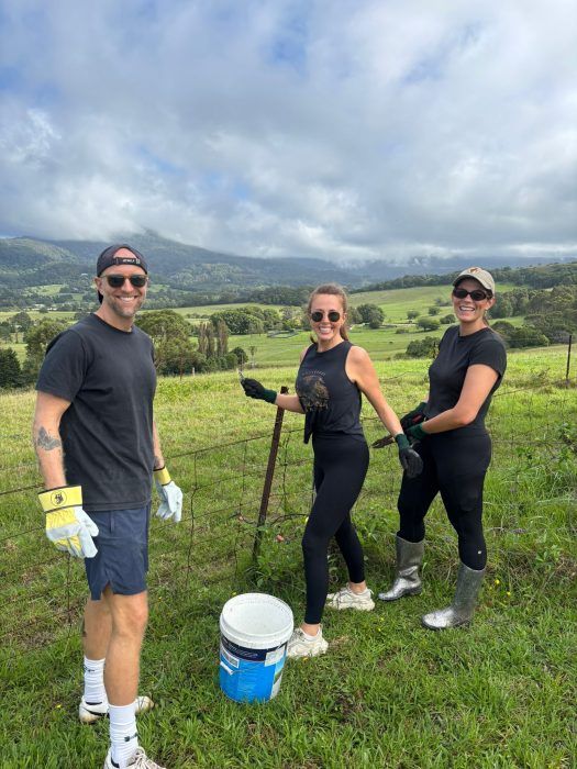 Three people in casual clothing and work gloves stand in a grassy field with a bucket, mountains in the background.