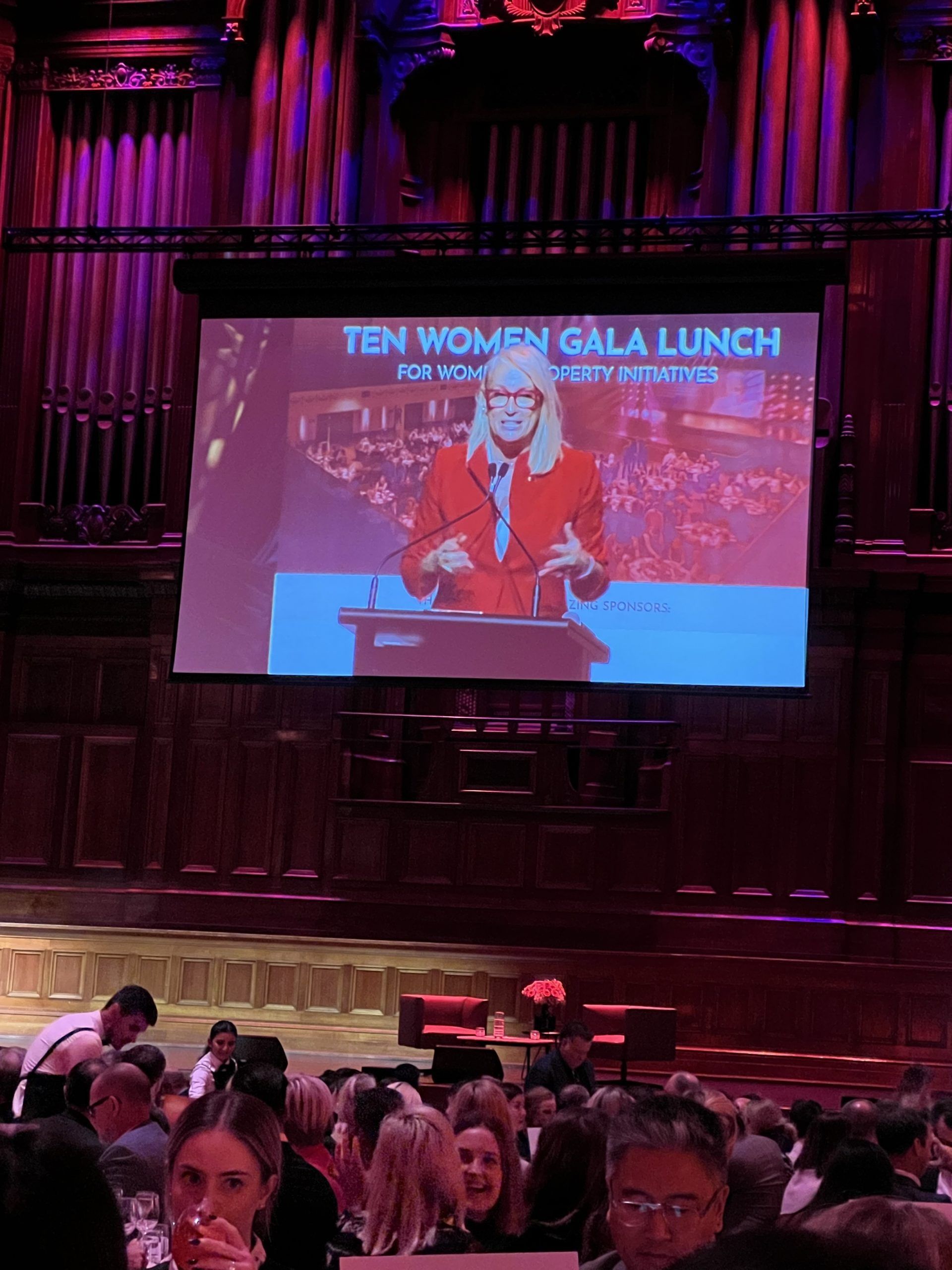A screen displays a speaker in a red blazer at a 
