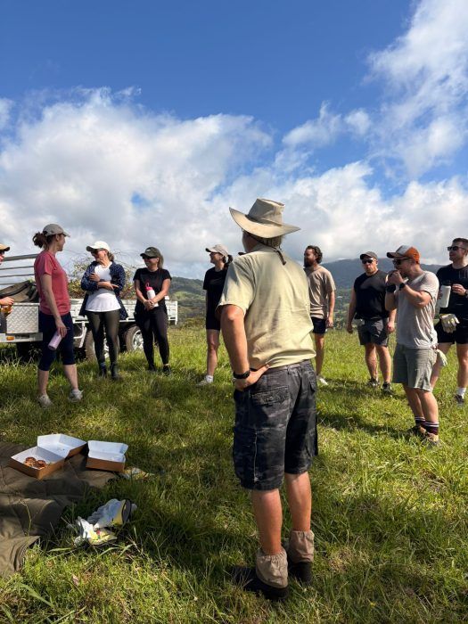 A group of people wearing casual clothing and hats stand outdoors in a grassy field during a bright, sunny day.