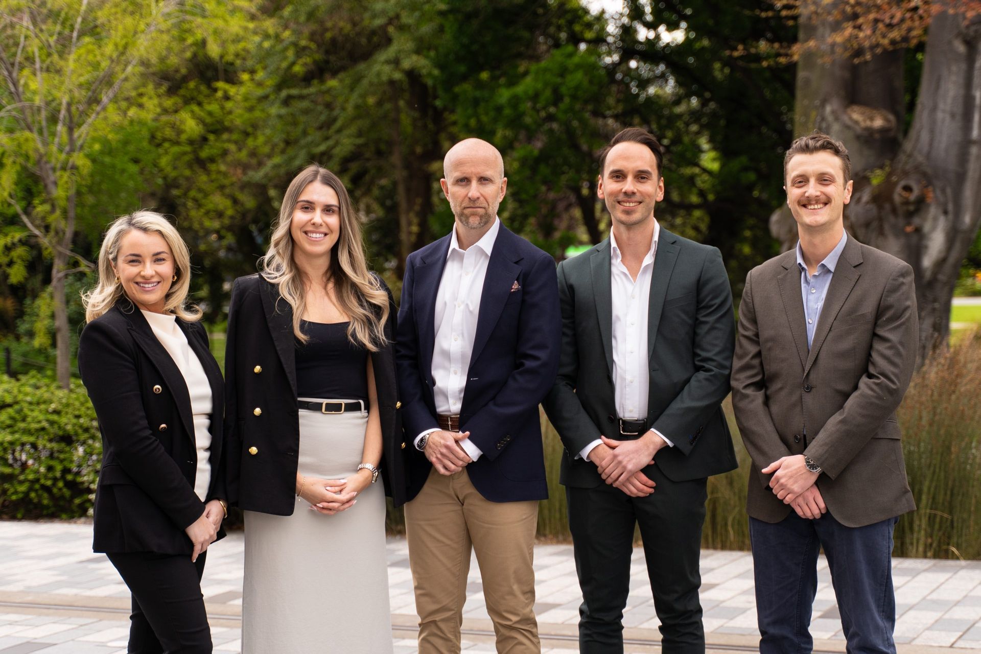 Five people in professional attire stand side-by-side posing for a photo in an outdoor park setting.