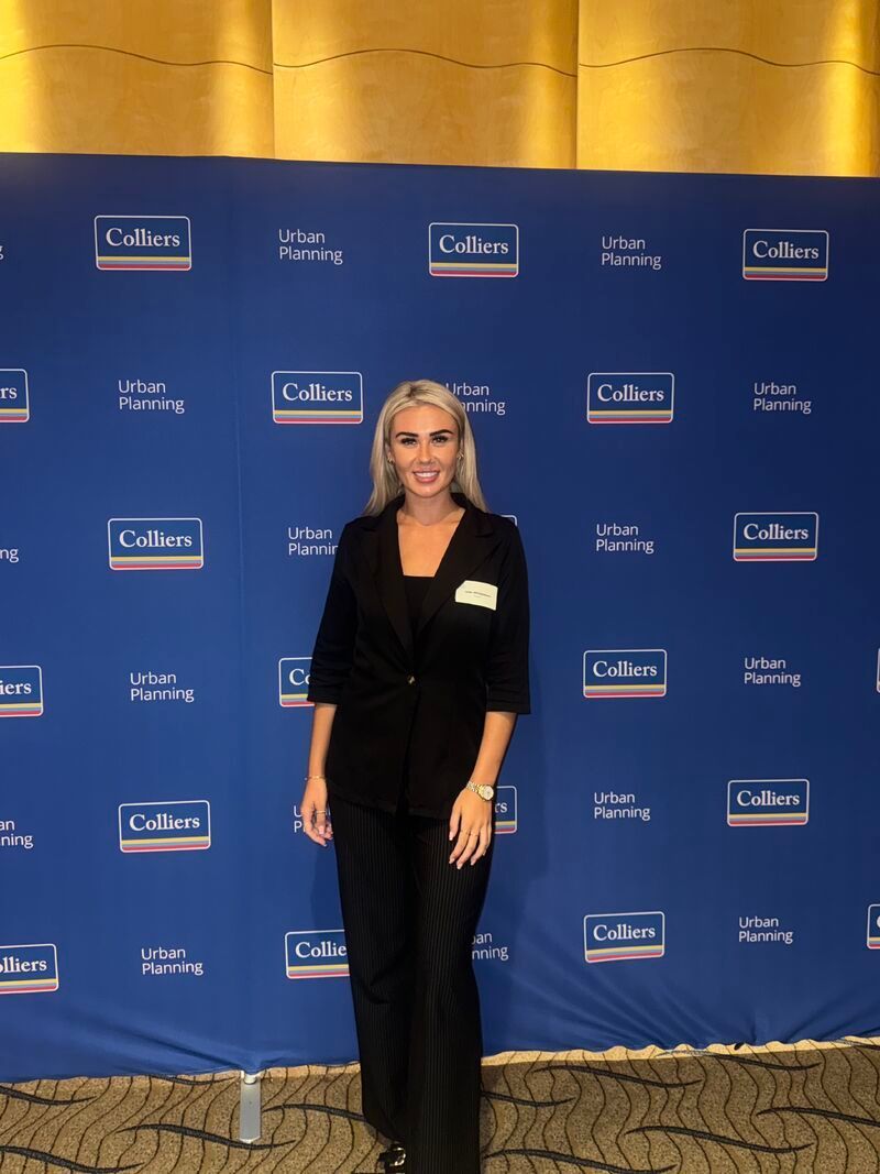A smiling person in a black blazer and trousers stands before a blue step-and-repeat backdrop featuring Colliers logos.