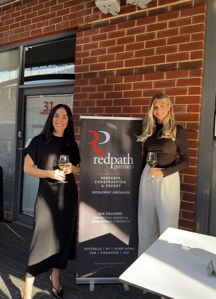 Two people holding wine glasses stand on either side of a Redpath Partners branded banner outside a brick building.