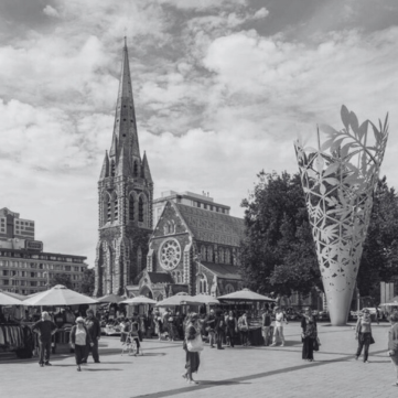 People browse a sunny outdoor market in Christchurch, New Zealand, beside the historic cathedral and a tall art sculpture.