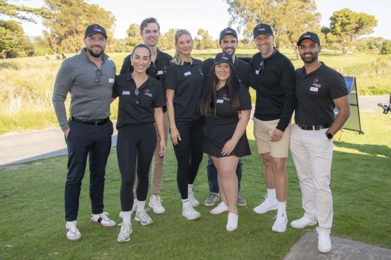 A group of eight smiling people in casual athletic wear and baseball caps pose outdoors on a sunny day.