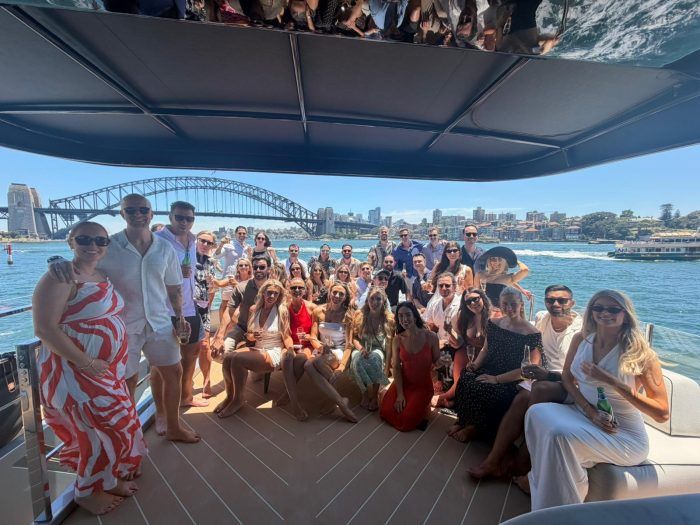 A group of people pose for a sunny boat cruise on Sydney Harbour with the Harbour Bridge in the background.