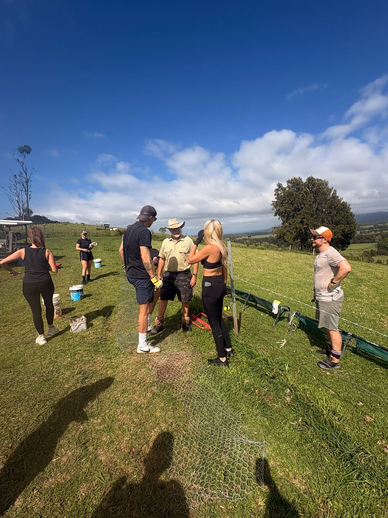 A group of people standing on a grassy hill under a blue sky, interacting near equipment setup in a rural field.