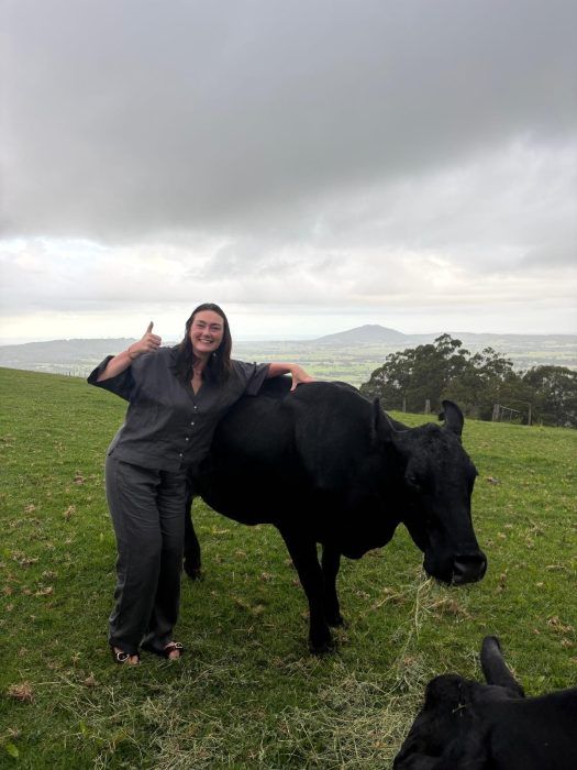 A smiling person in dark clothing stands in a grassy field, leaning on a black cow while giving a thumbs up.