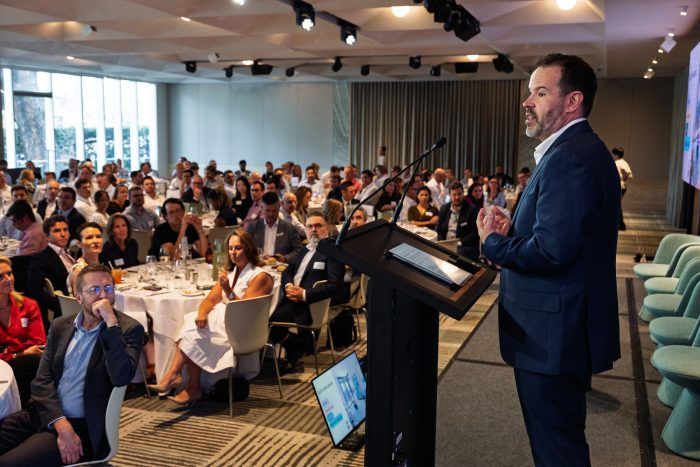 A man in a suit speaks at a podium to an audience seated at tables in a large, brightly lit conference hall.