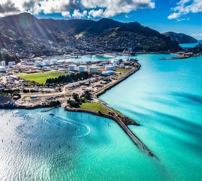 Aerial view of Lyttelton Harbour, New Zealand, showing a coastal industrial port with storage tanks and bright turquoise water.