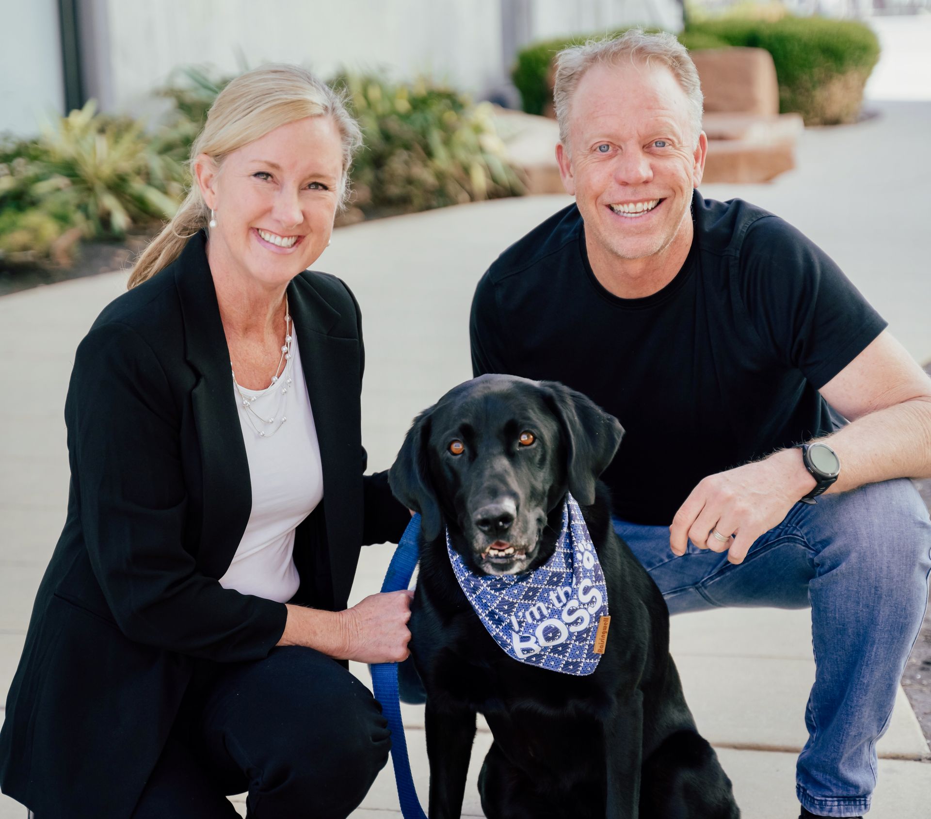 A man and woman are posing for a picture with a black dog wearing a bandana that says i 'm the boss