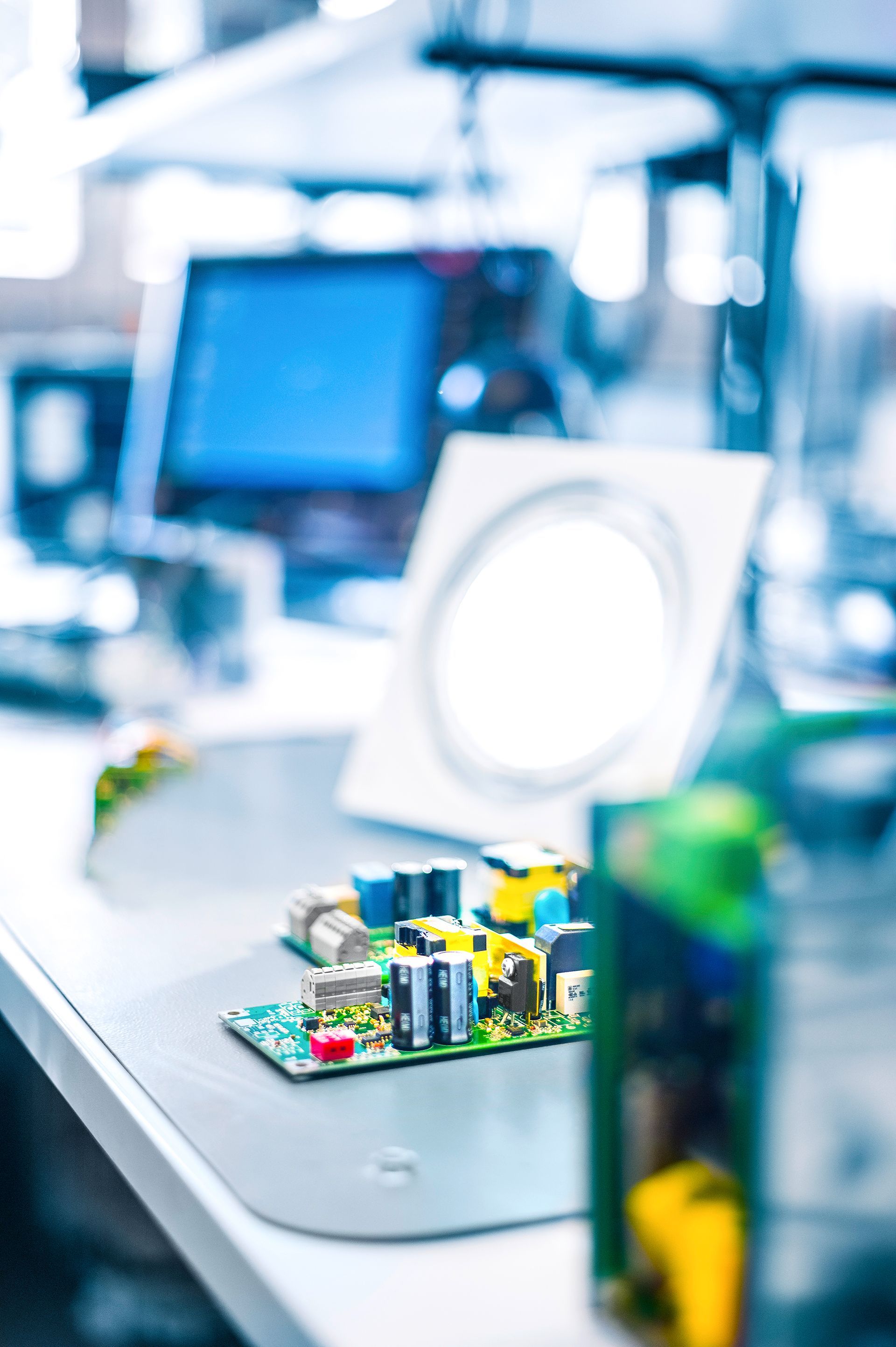 Leiterplatte mit verschiedenen elektronischen Komponenten auf einem Schreibtisch in einer Laborumgebung, mit einem Computermonitor und einer Inspektionslampe im Hintergrund.