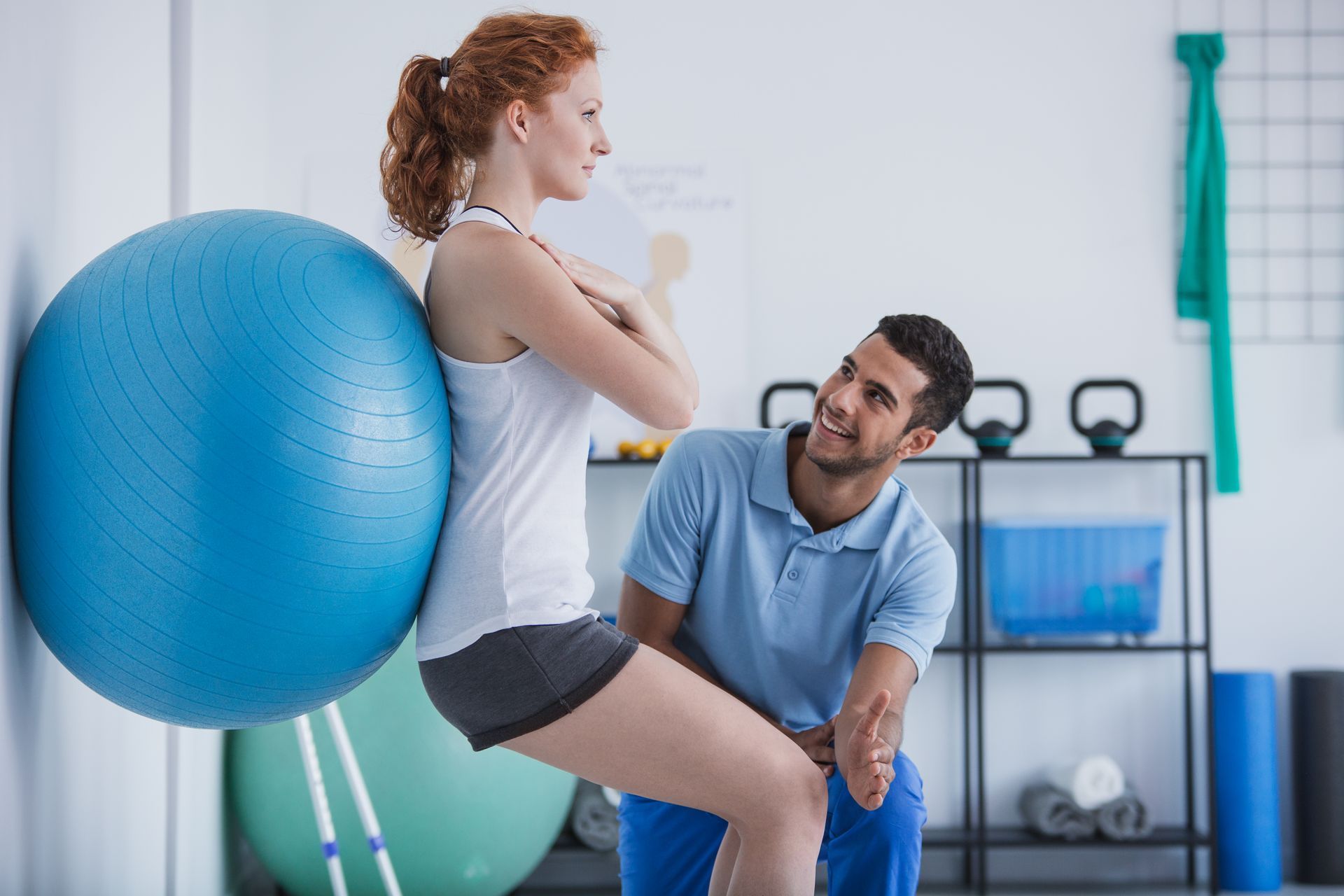 A man is helping a woman with a pilates ball in a gym.