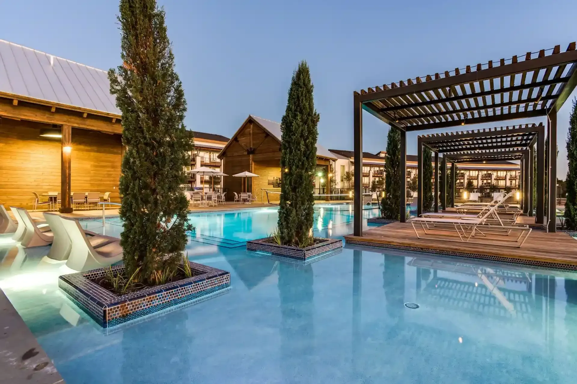 Pool with a pergola, trees, and lounge chairs in an outdoor setting at dusk at The Boat House, offers apartments and townhomes in The Colony, TX.