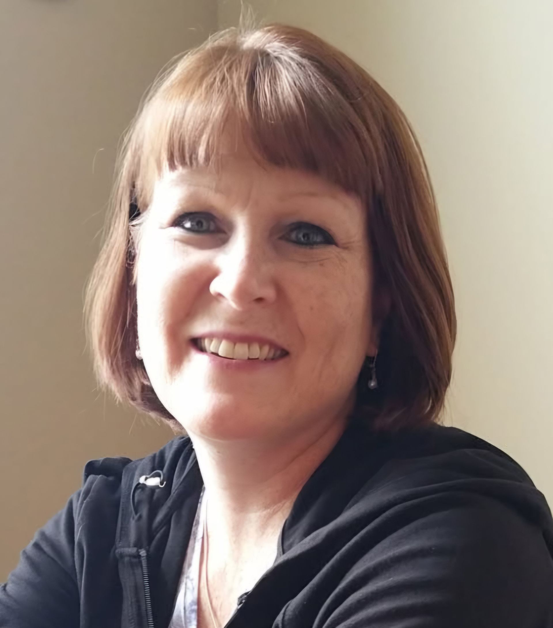 Woman with reddish-brown hair smiling, wearing a black jacket, against a neutral background.
