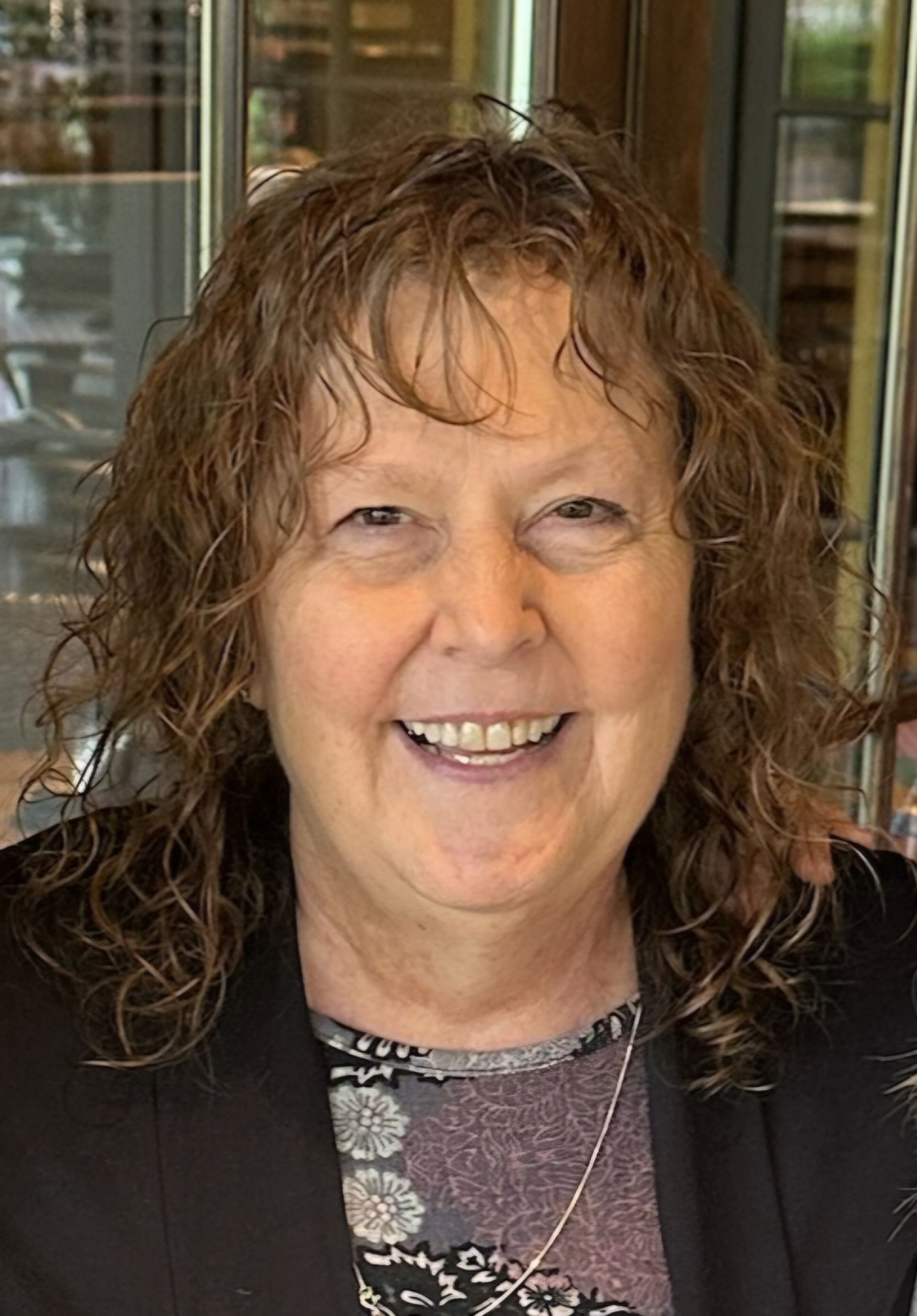 Woman with brown curly hair, smiling, wearing a dark jacket, sitting indoors.