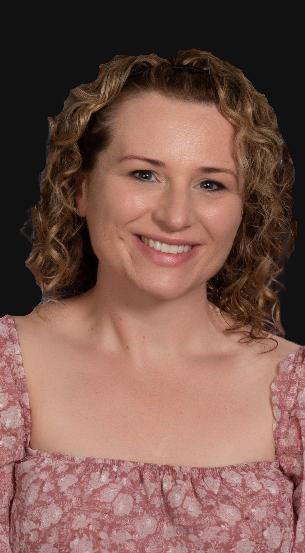 Woman with curly blonde hair smiles at the camera, wearing a pink floral top. Dark background.