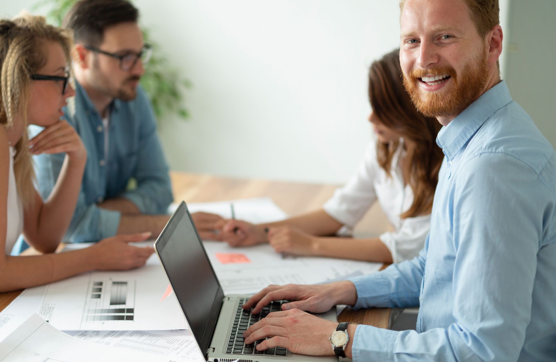 Man with red beard smiles, typing on laptop at a table with colleagues looking at papers.