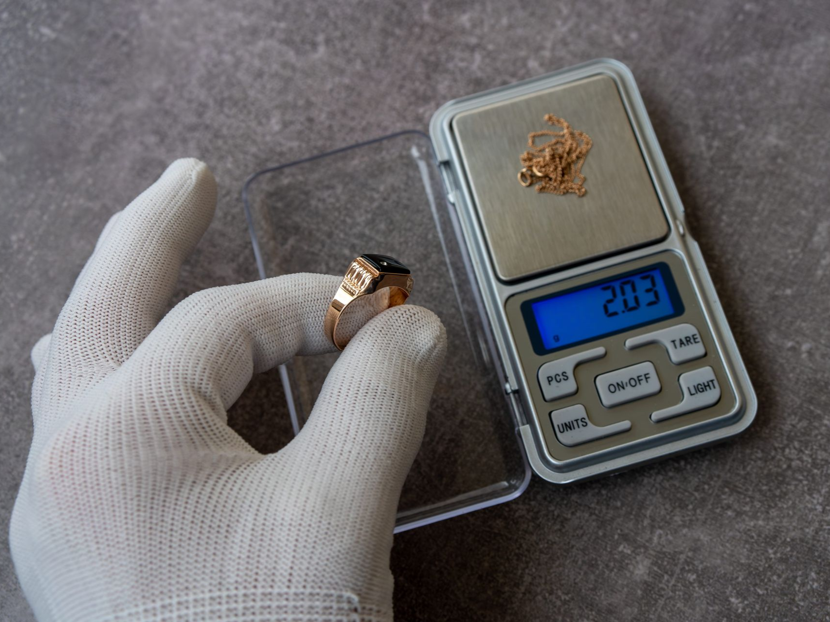 A gloved hand holds a gold ring near a digital scale measuring a small piece of gold jewelry.