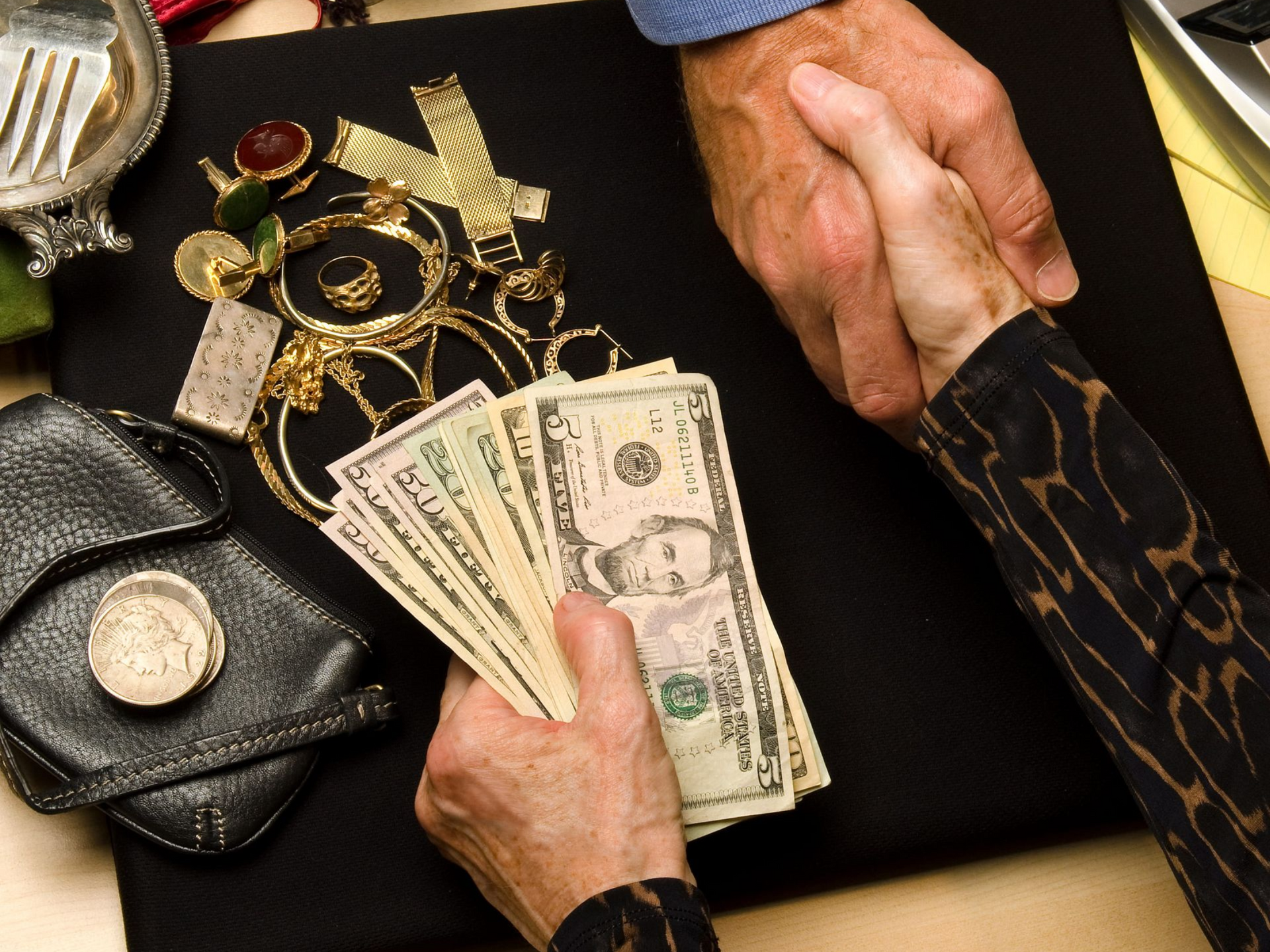 Hands shaking over a collection of gold jewelry and a stack of US dollar bills on a black surface.