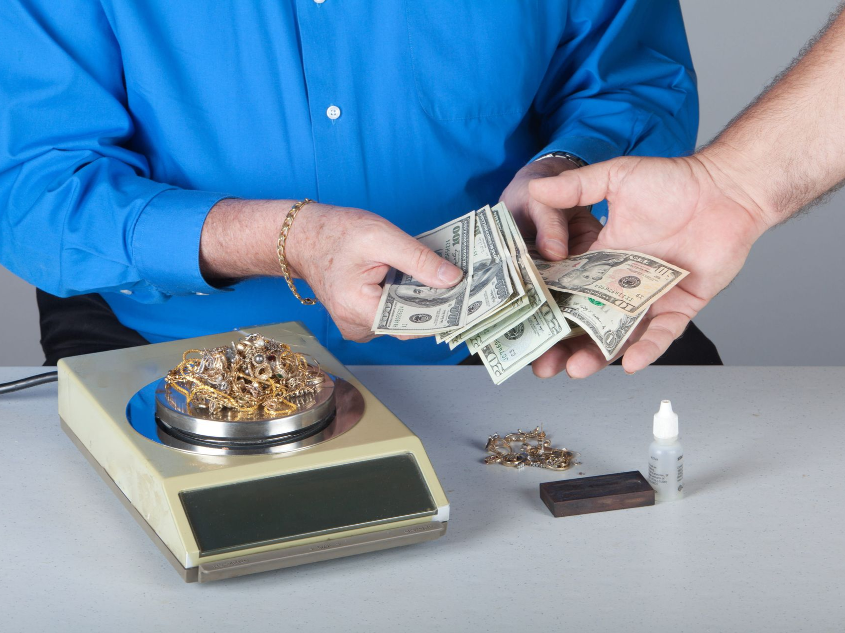 A person in a blue shirt holds cash for gold jewelry weighed on a scale.