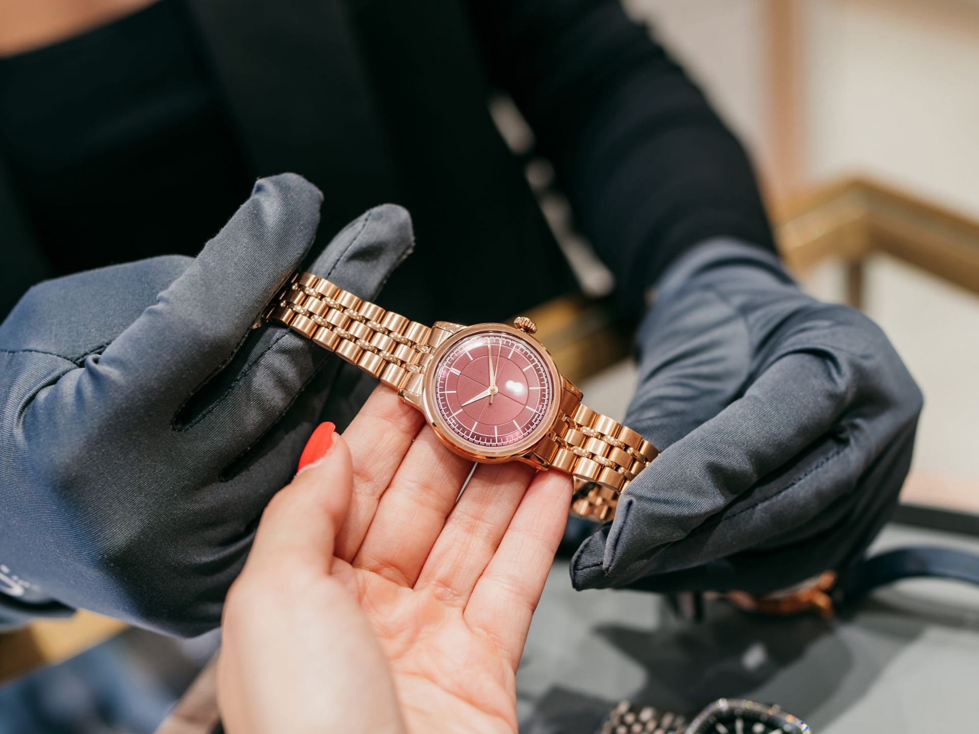 Hands in dark gloves hold a rose gold watch with a dark red face above a person's open palm.