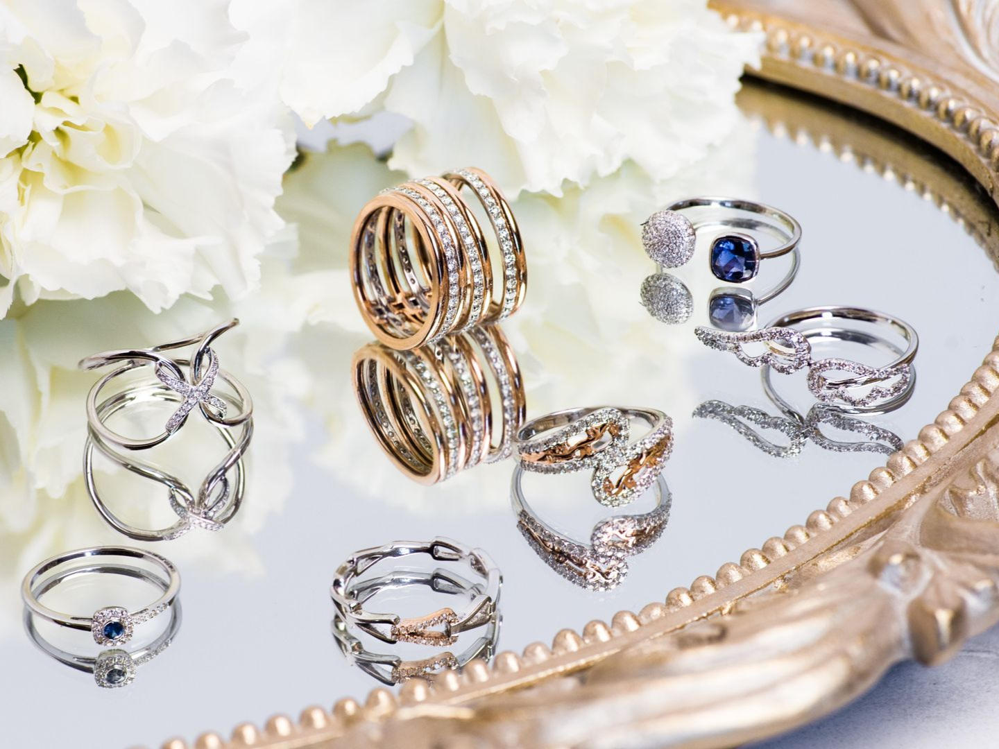 A collection of rings arranged on a reflective mirrored tray next to white flowers.