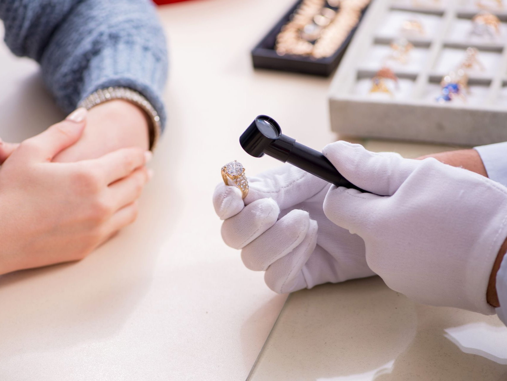 A jeweler in white gloves uses a loupe to inspect a diamond ring held for a customer at a jewelry store counter.