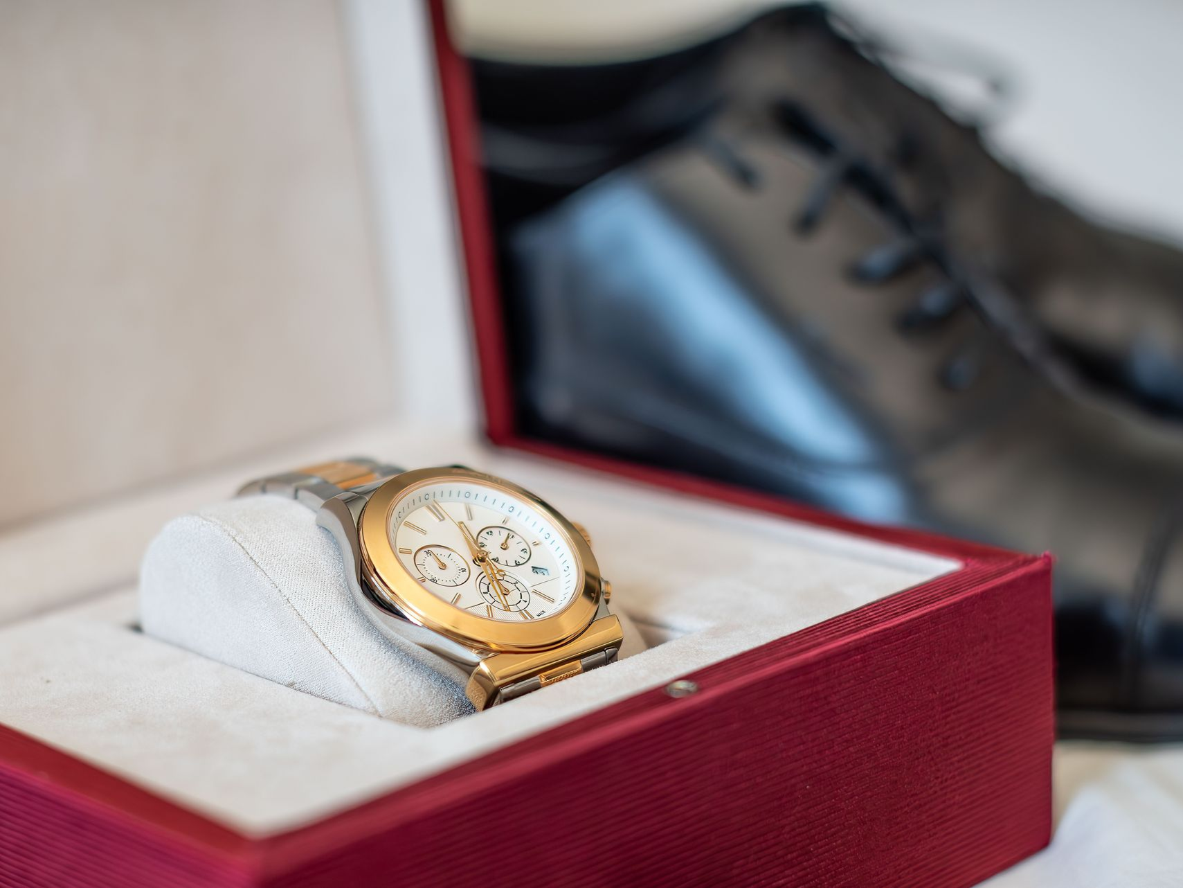 A silver and gold chronograph watch displayed in an open red jewelry box next to a pair of black leather dress shoes.