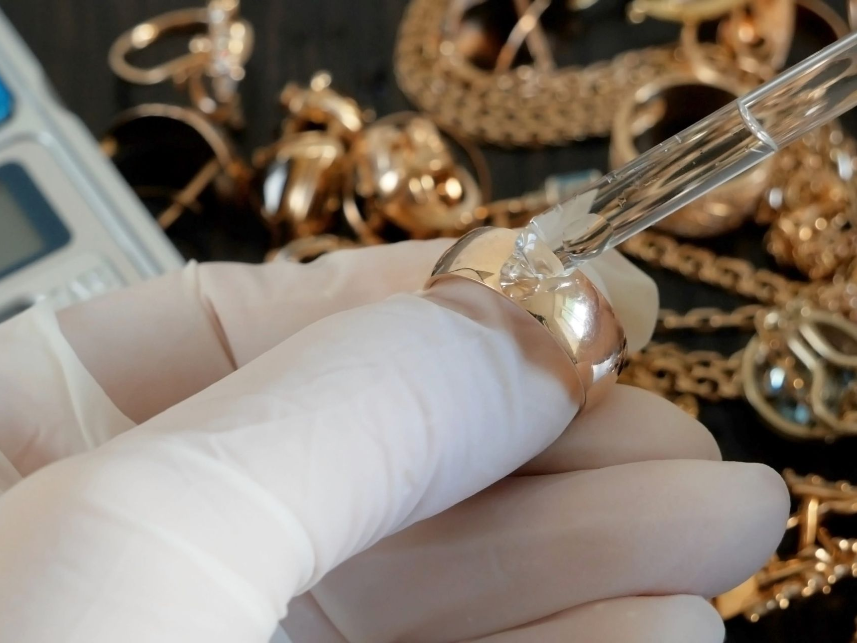 A gloved hand uses a dropper to apply acid testing solution to a gold ring, surrounded by jewelry on a dark surface.