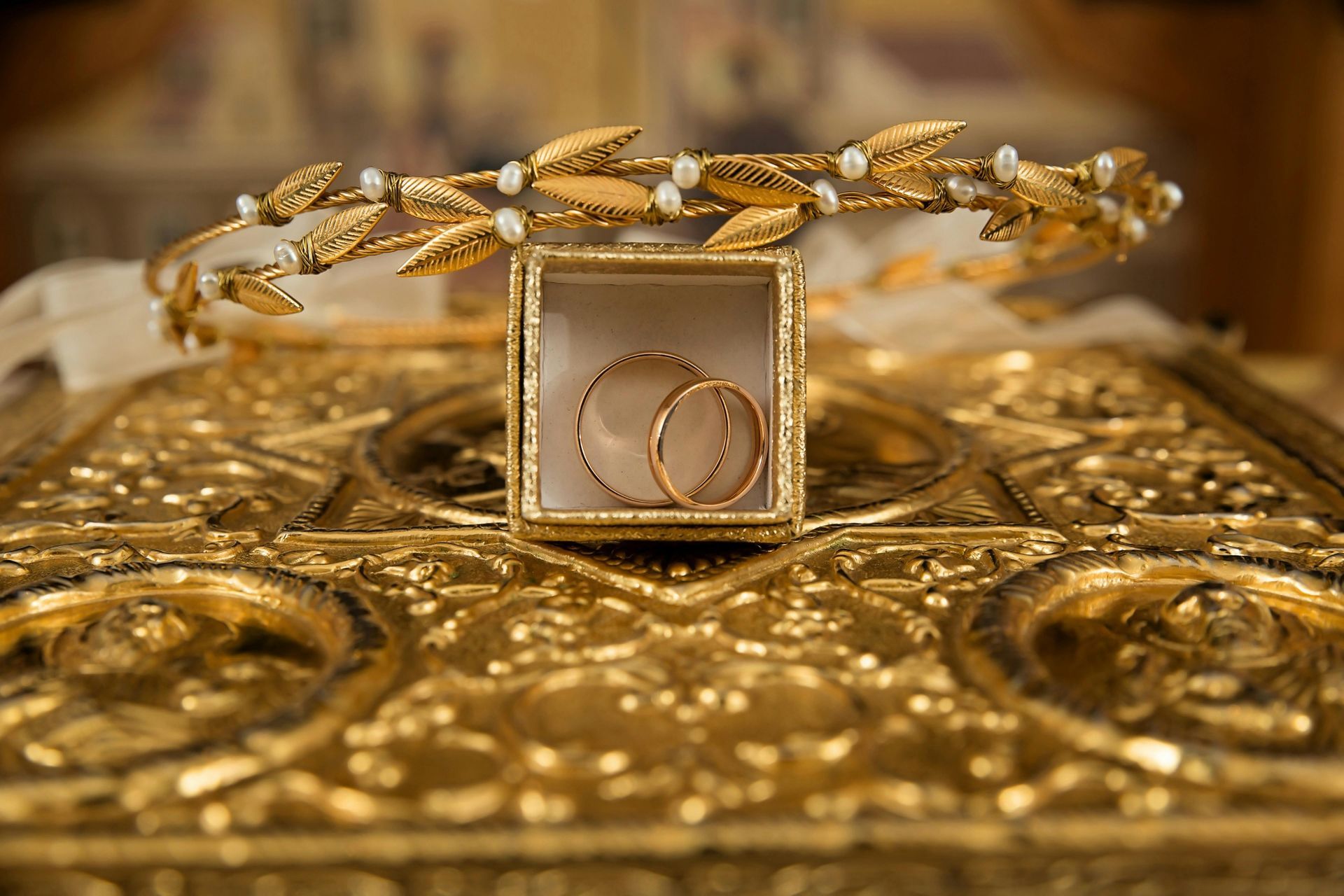 Two gold wedding rings in a small square glass box sit atop an ornate gold surface, below a decorative gold leaf crown.