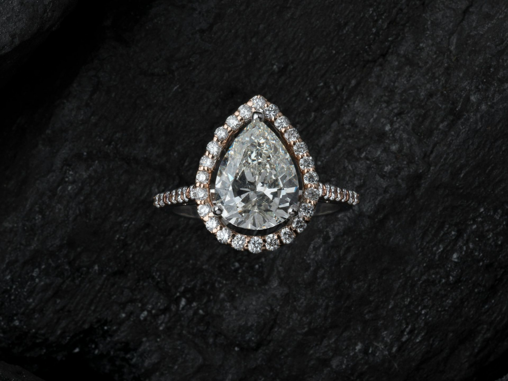 A pear-cut diamond ring with a halo setting and a pave band, centered on a dark, textured charcoal background.