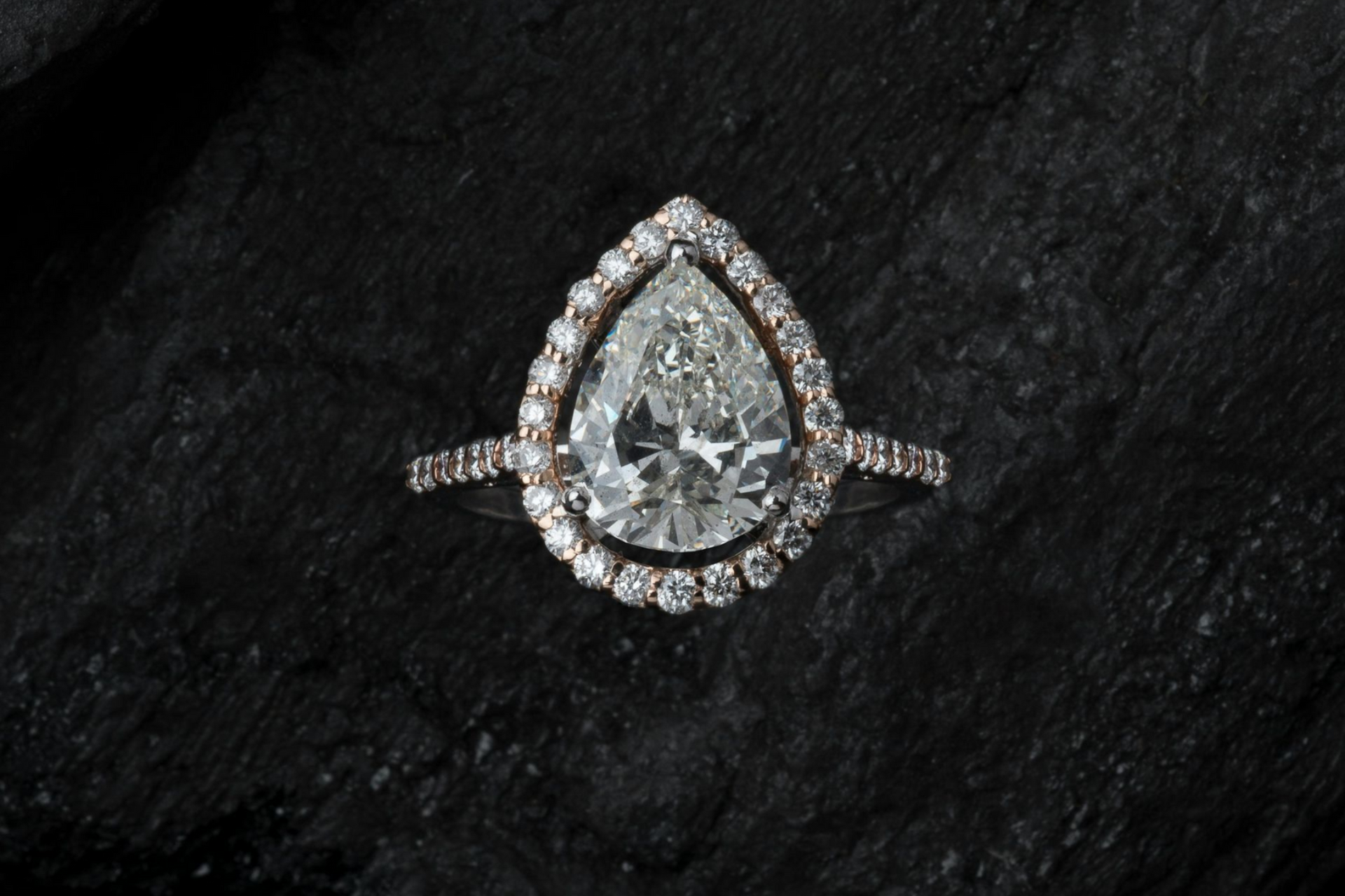 A pear-cut diamond engagement ring with a halo of smaller diamonds, set against a dark, textured charcoal background.