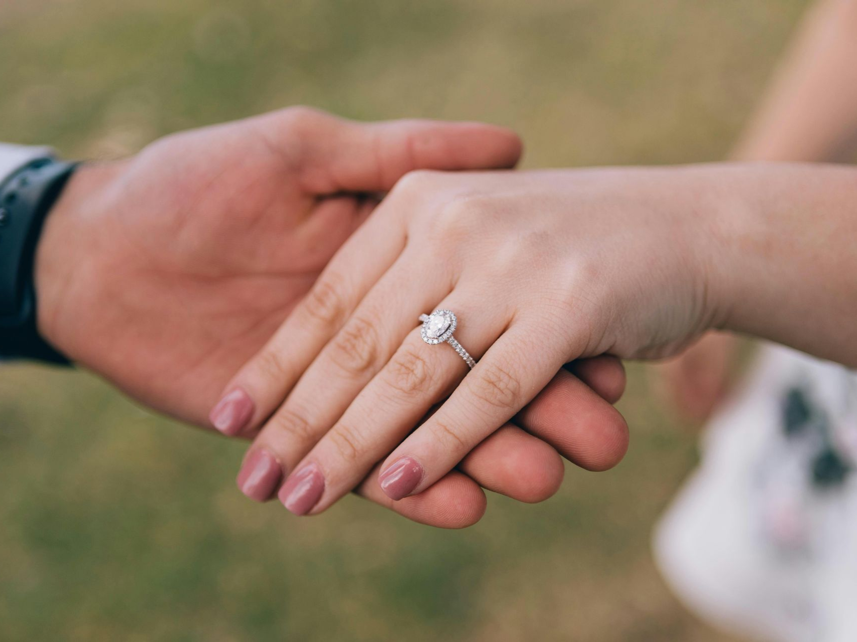 A person’s hand rests in another’s, showcasing a silver engagement ring with a central pear-shaped diamond outdoors.