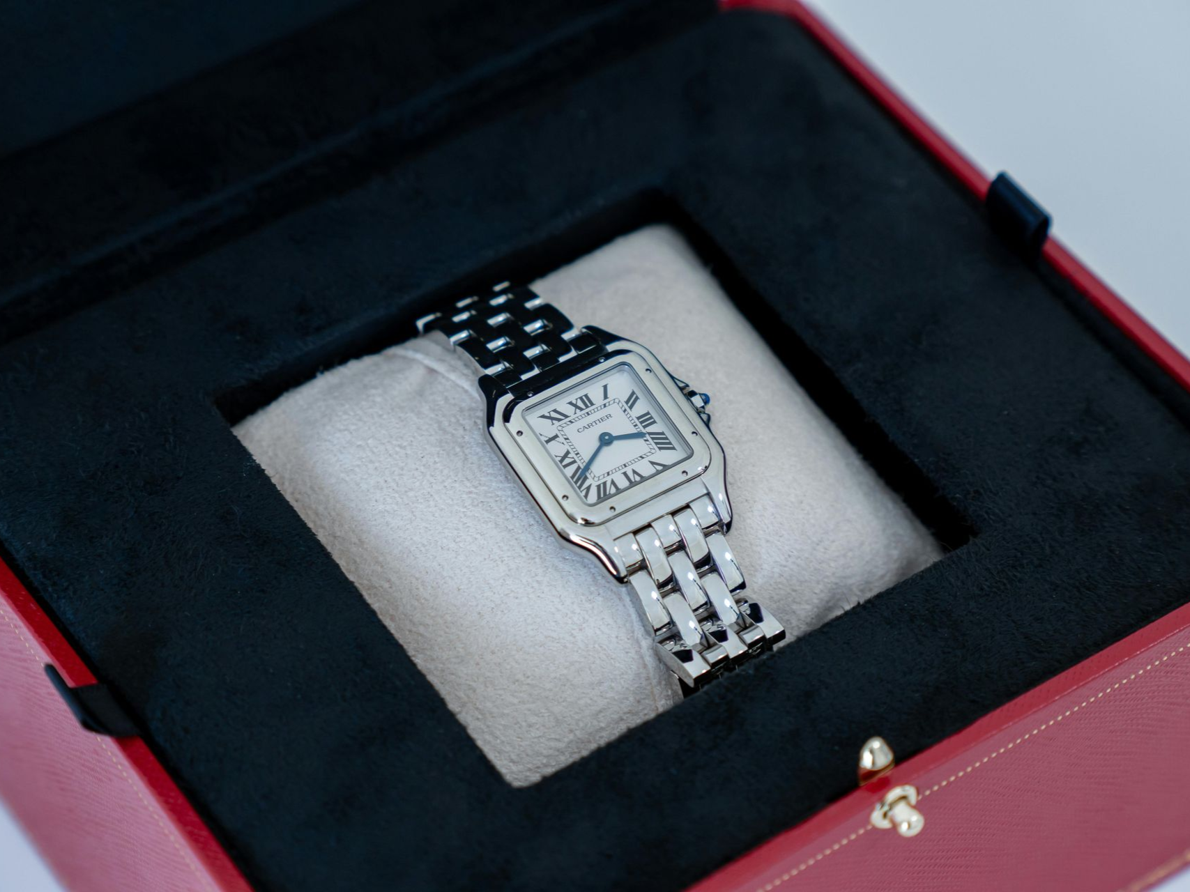 A silver Cartier Panthère watch with a link bracelet, resting on a beige velvet pillow inside a red presentation box.
