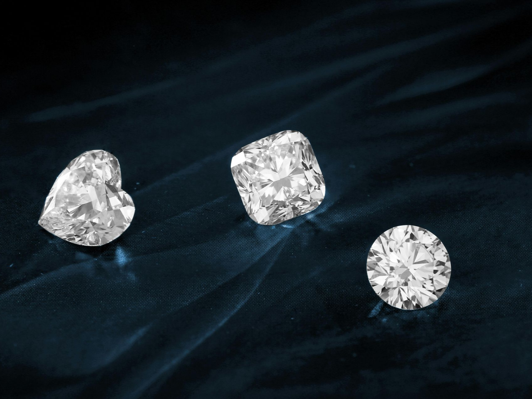Three loose diamonds—heart, cushion, and round cut—rest on a dark blue velvet surface.