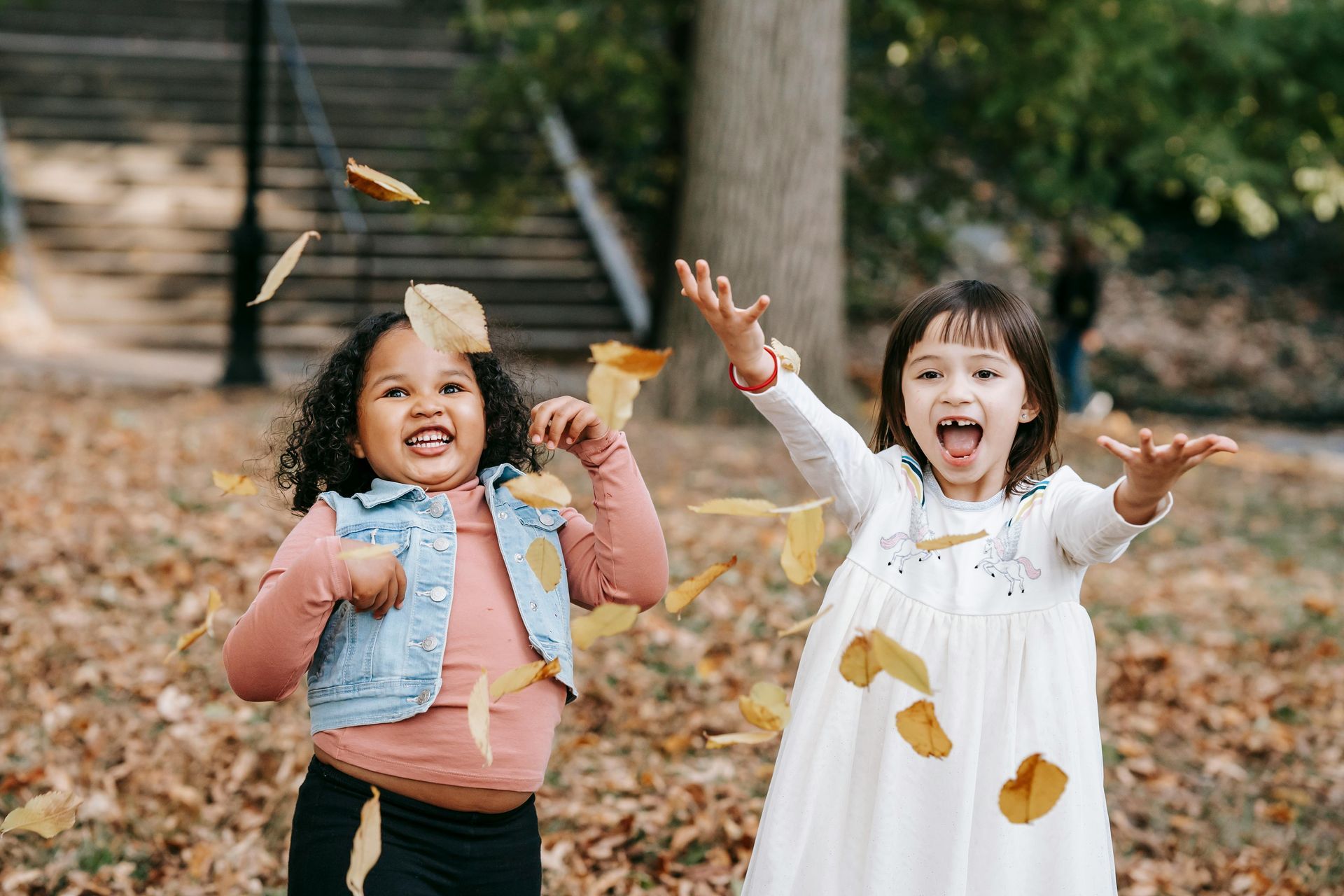 Two little girls are throwing leaves in the air in a park.