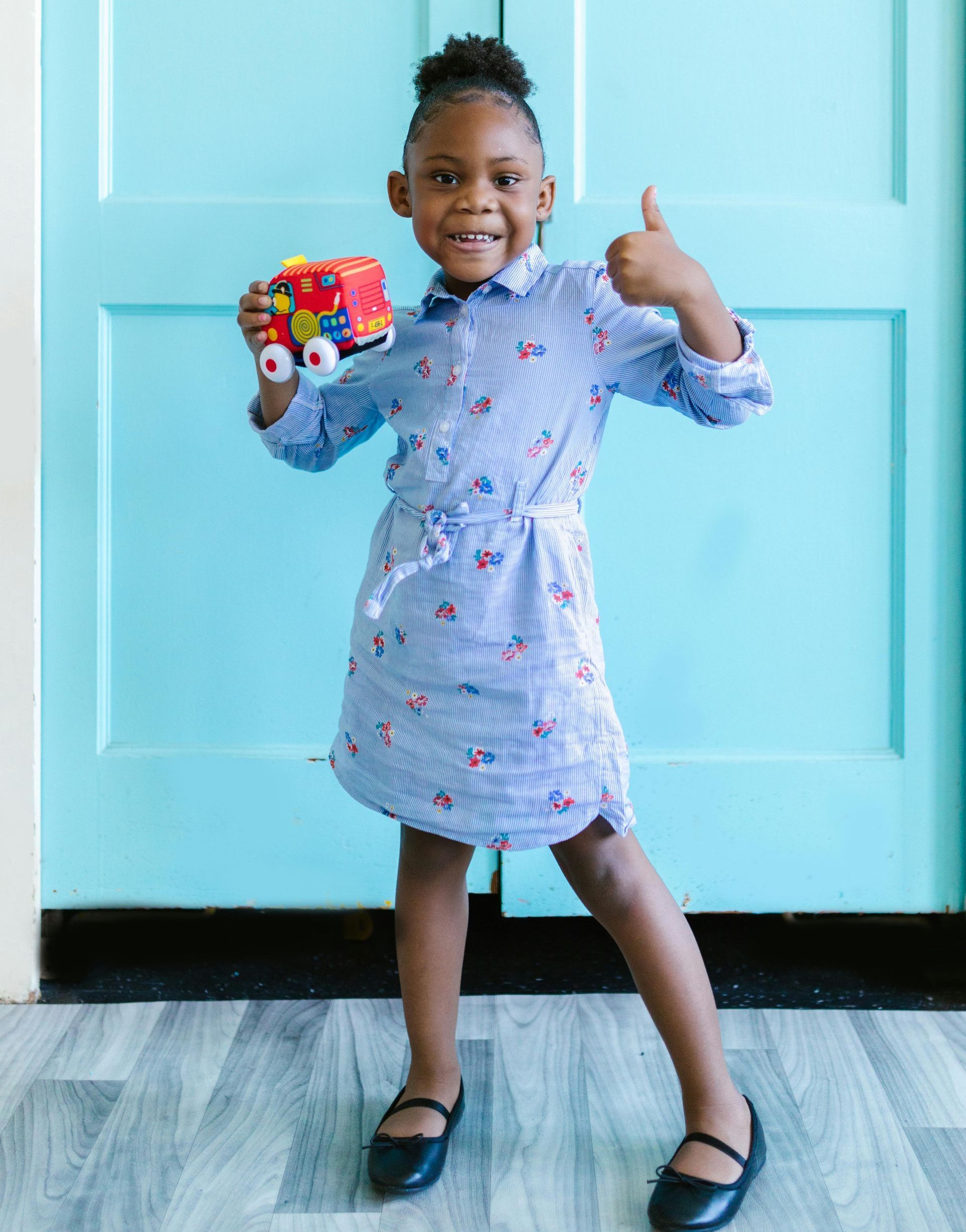 A little girl in a blue dress is holding a toy and giving a thumbs up.