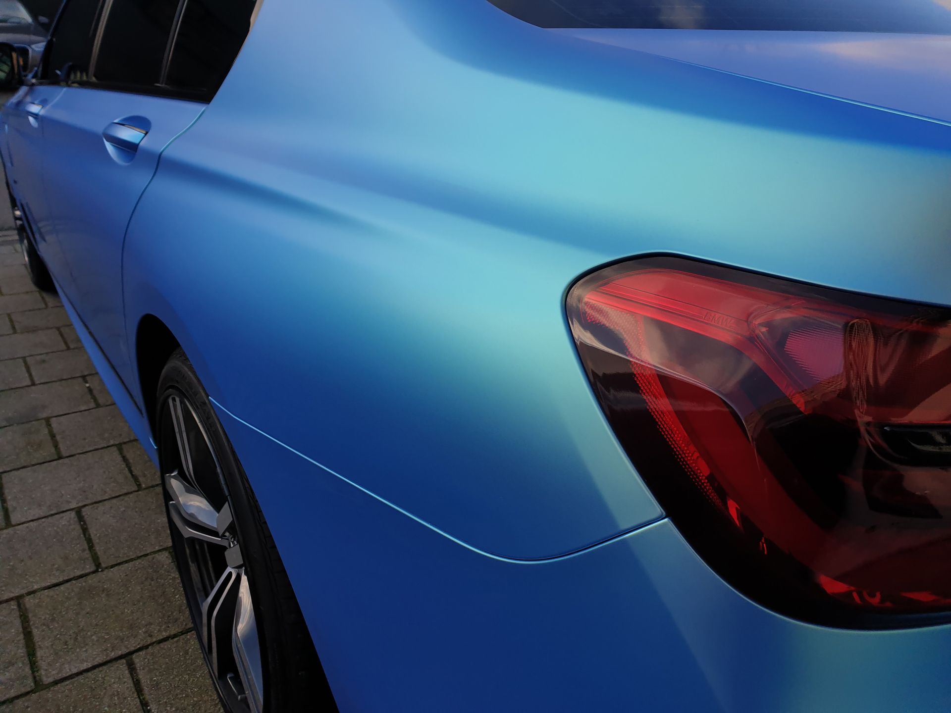 A blue car with a red tail light is parked on a sidewalk.