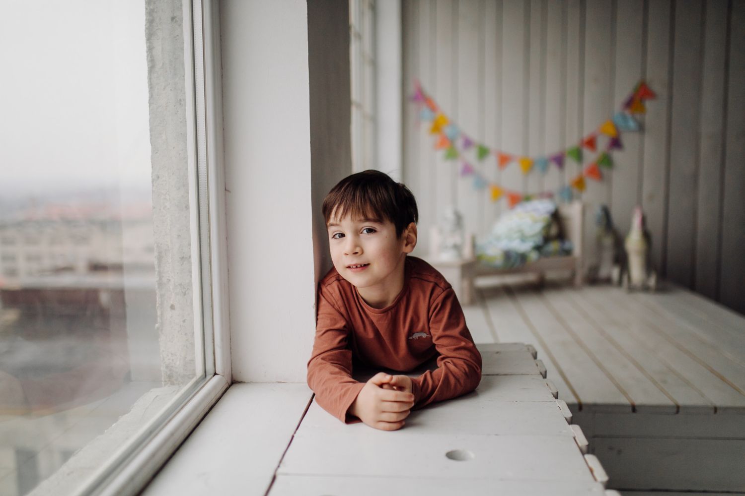 An autistic boy is laying on a window sill looking out the window.