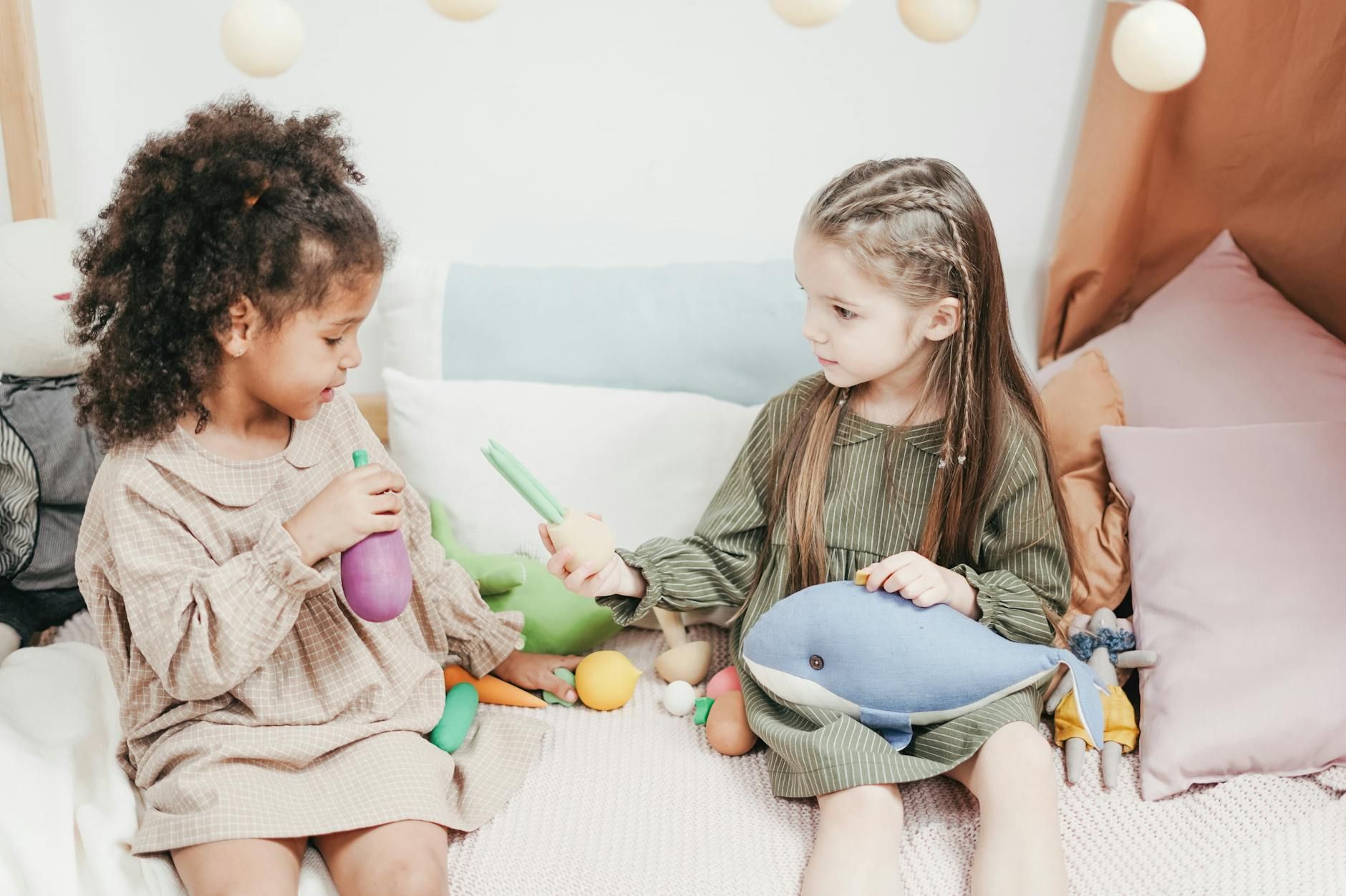 Two autistic girls sharing toy vegetables and a plush dolphin on a bed during in-home ABA therapy.