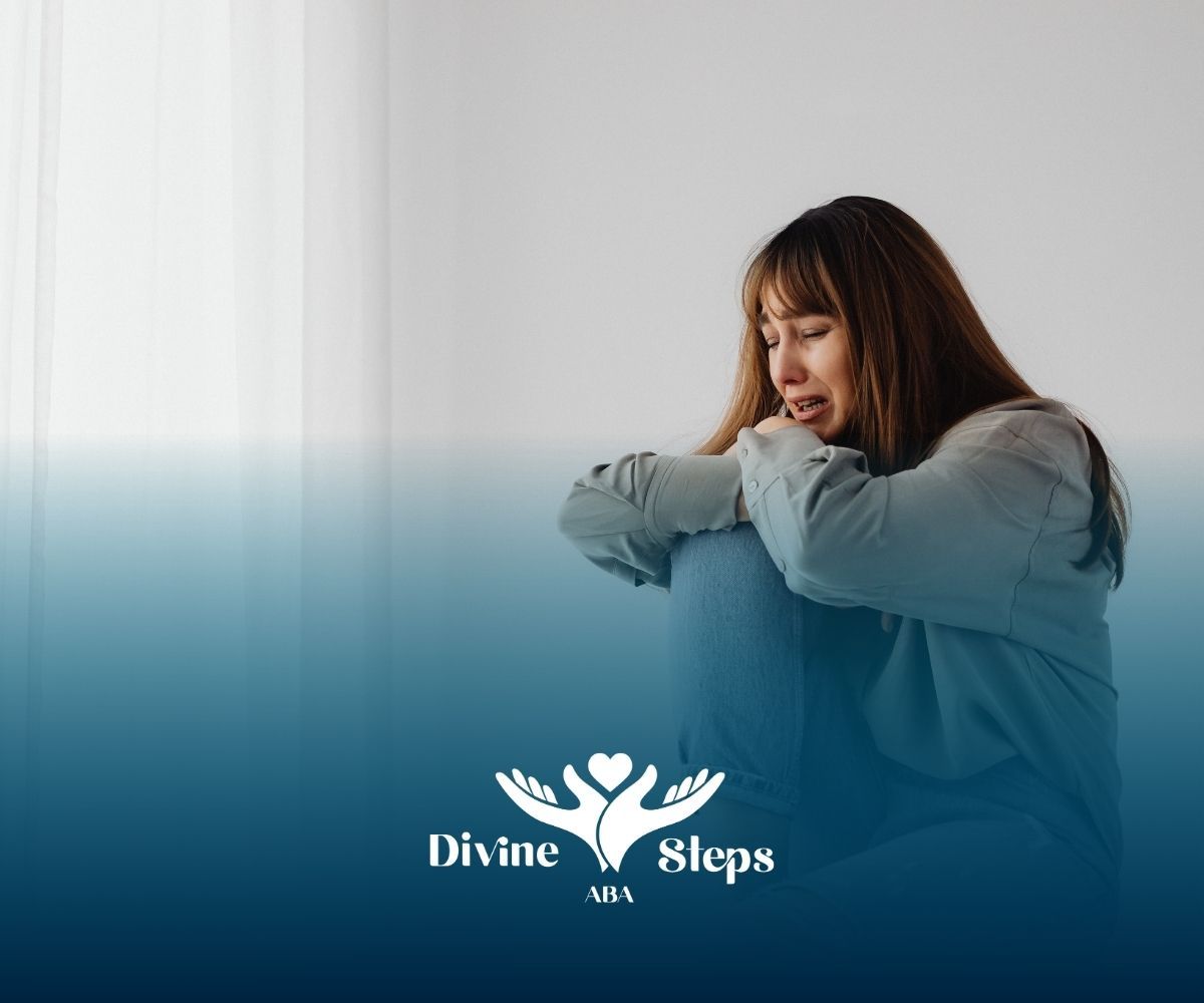 Woman in a distressed pose, hugging knees, in a white room. Divine Steps USA logo at bottom.