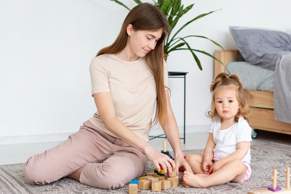 Woman and child playing with wooden blocks on a rug. The child looks at the camera.