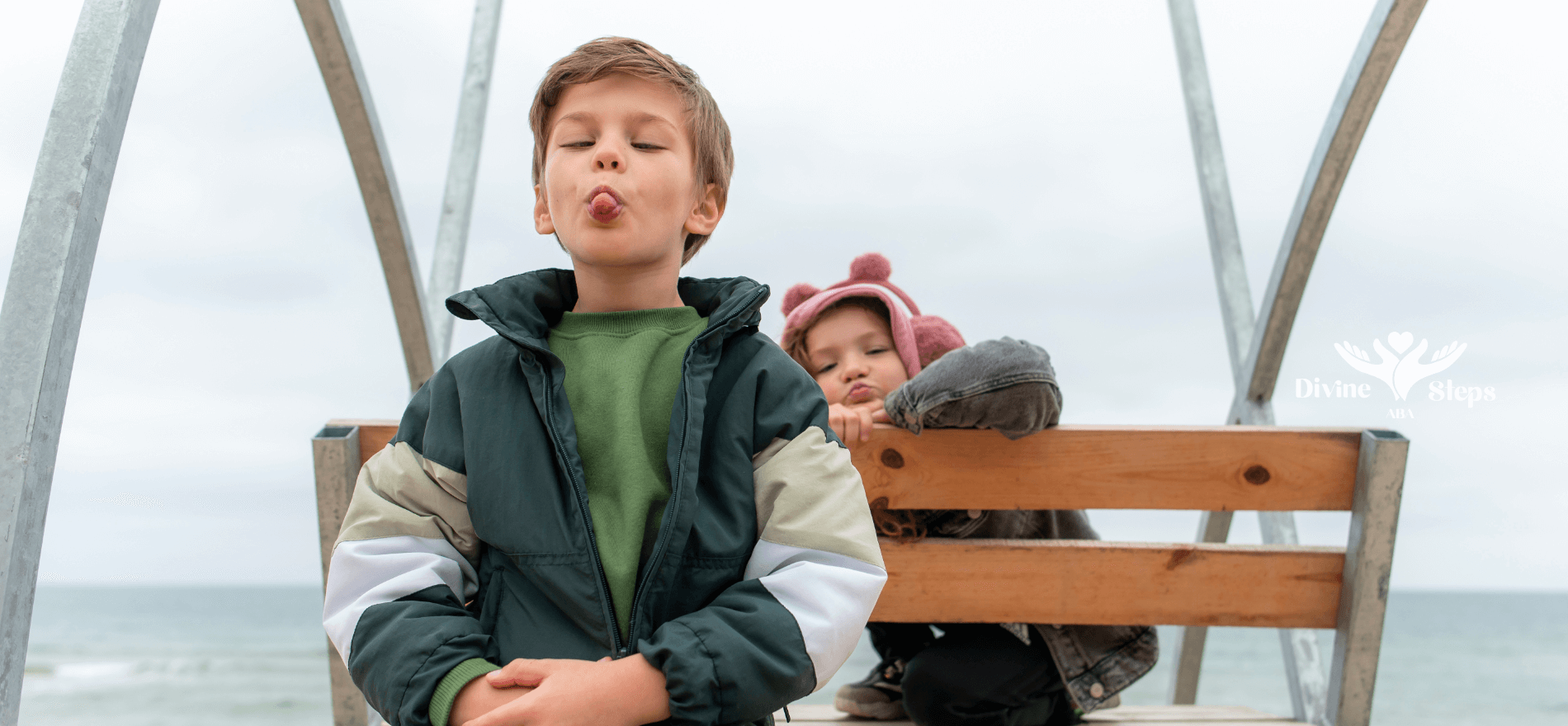An autistic kid is sitting on a wooden bench by the beach, making a playful face while another child