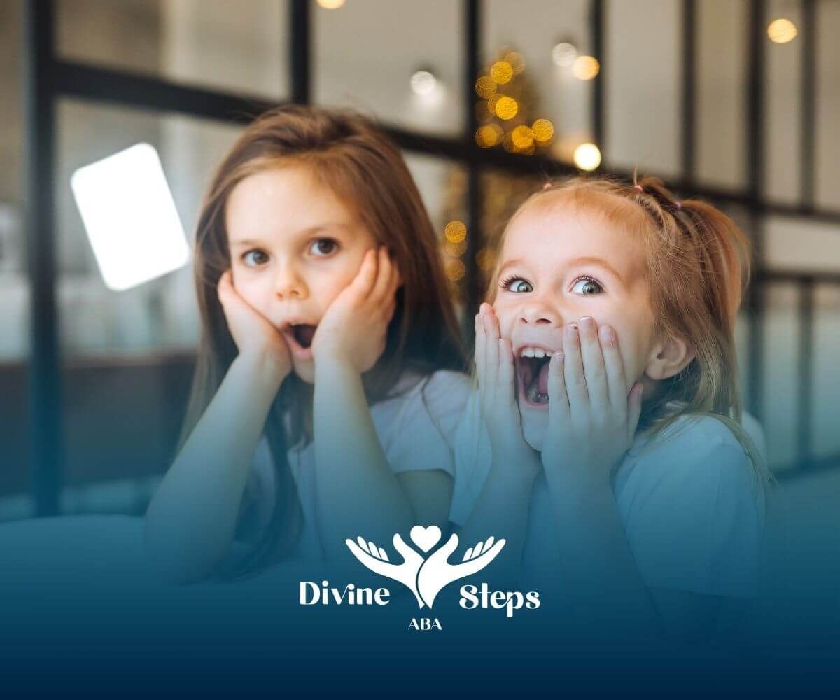 Two autistic girls with hands on cheeks showing surprise and excitement, sitting at a table indoors.