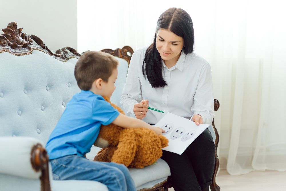 Woman pointing to a diagram, talking with a child holding a teddy bear on a blue couch.