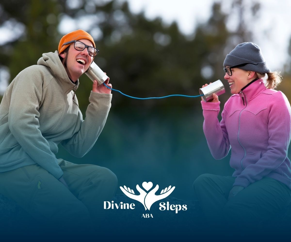 Two people laughing, using tin can phones outdoors. One wears an orange hat, the other a grey hat and pink jacket.