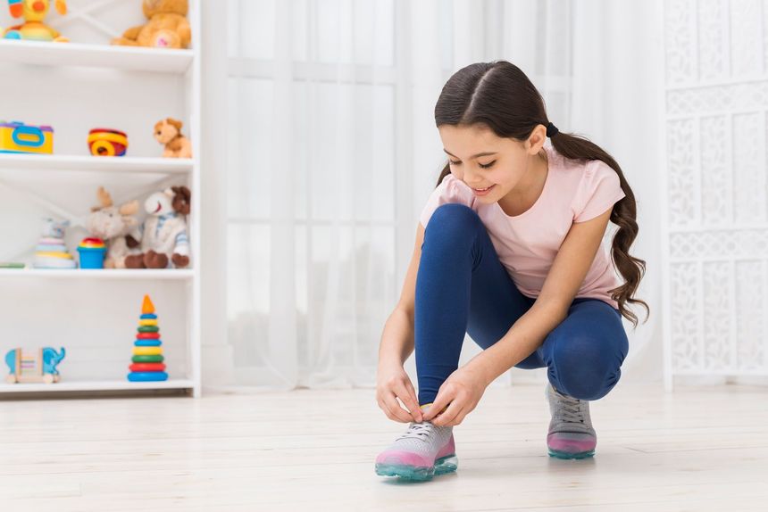 Girl in a pink shirt and blue leggings tying her shoelaces in a brightly lit room with a toy shelf.