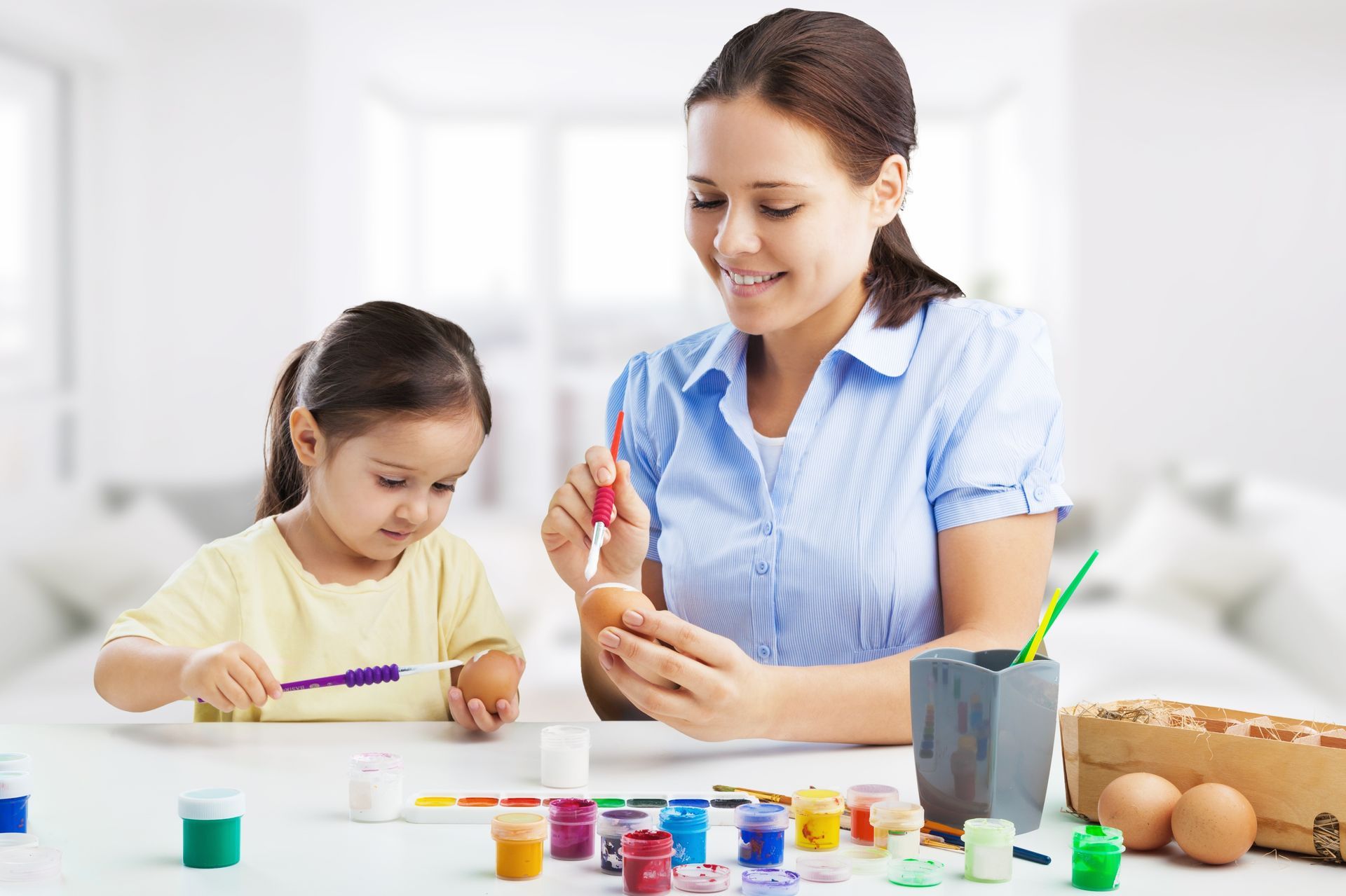Woman and child painting Easter eggs at a table with paint and brushes.